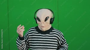 Ufo Alien mask with human clothes dance and listen music song with headphones and green wall in background - concept of earth planet and extraterrestrial in living between us